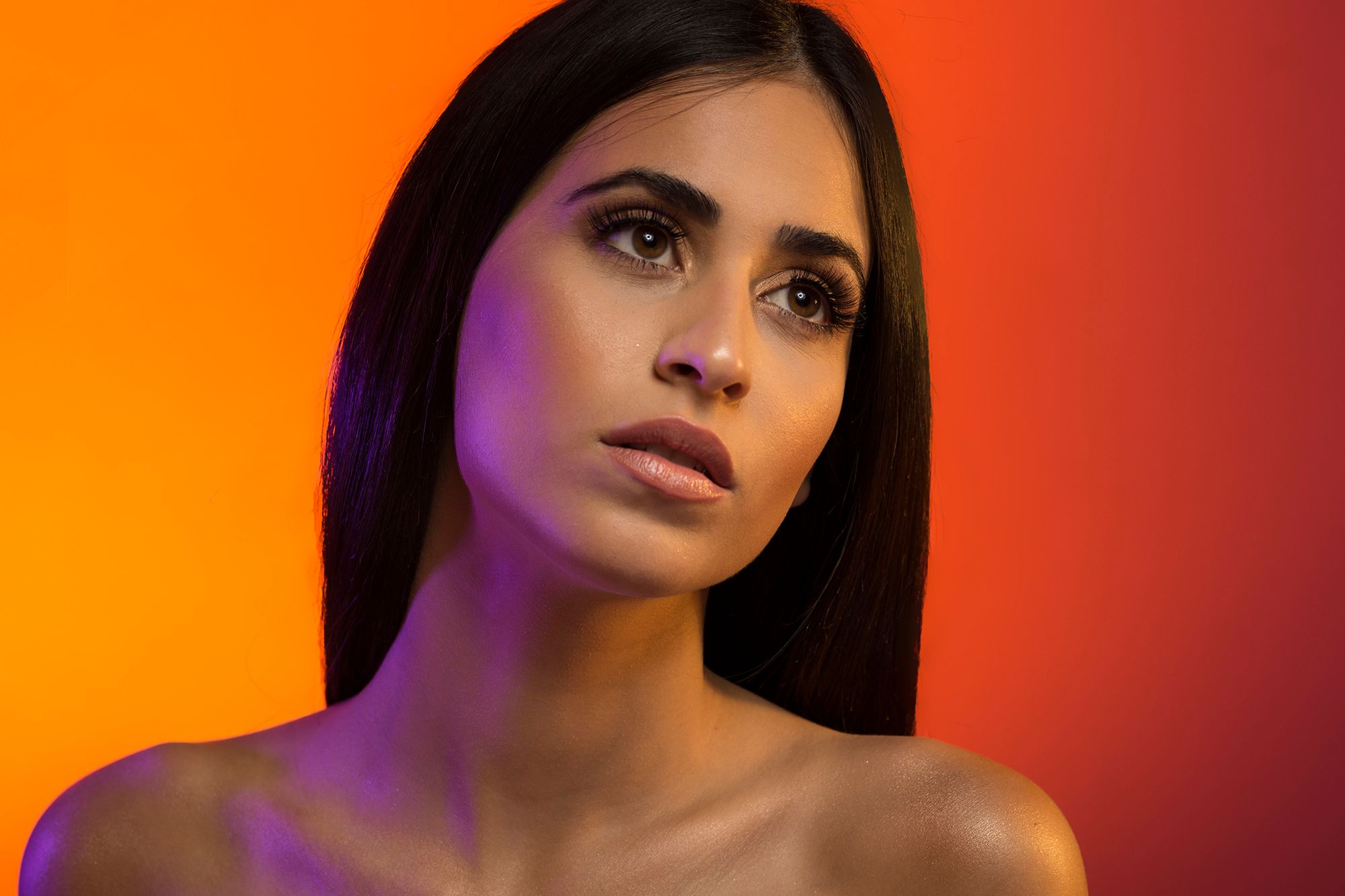 Studio beauty portrait with dramatic orange and red lighting against dark backdrop with purple accent illumination.