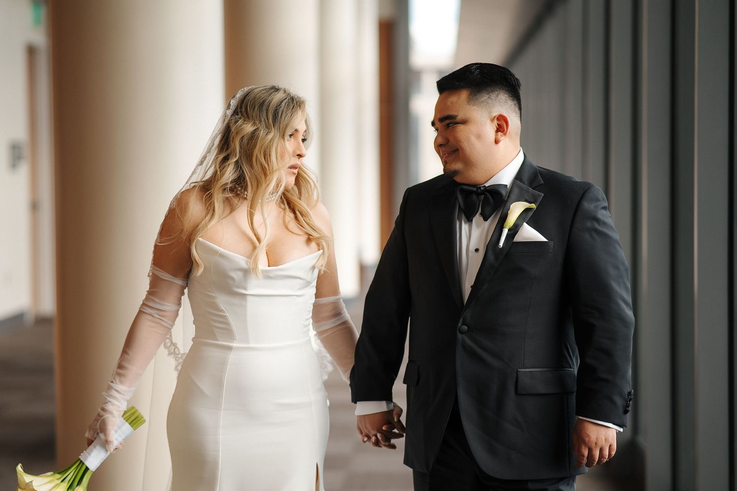 A bride in a white strapless gown and groom in a black tuxedo gaze at each other while holding hands.