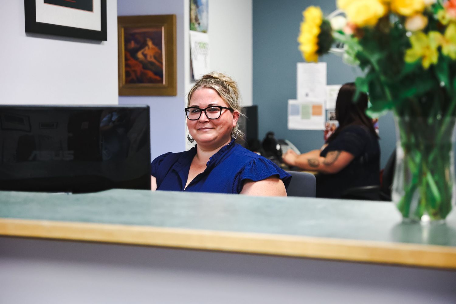 Business colleagues in glasses review documents while sitting at long conference table with laptops and yellow flowers.