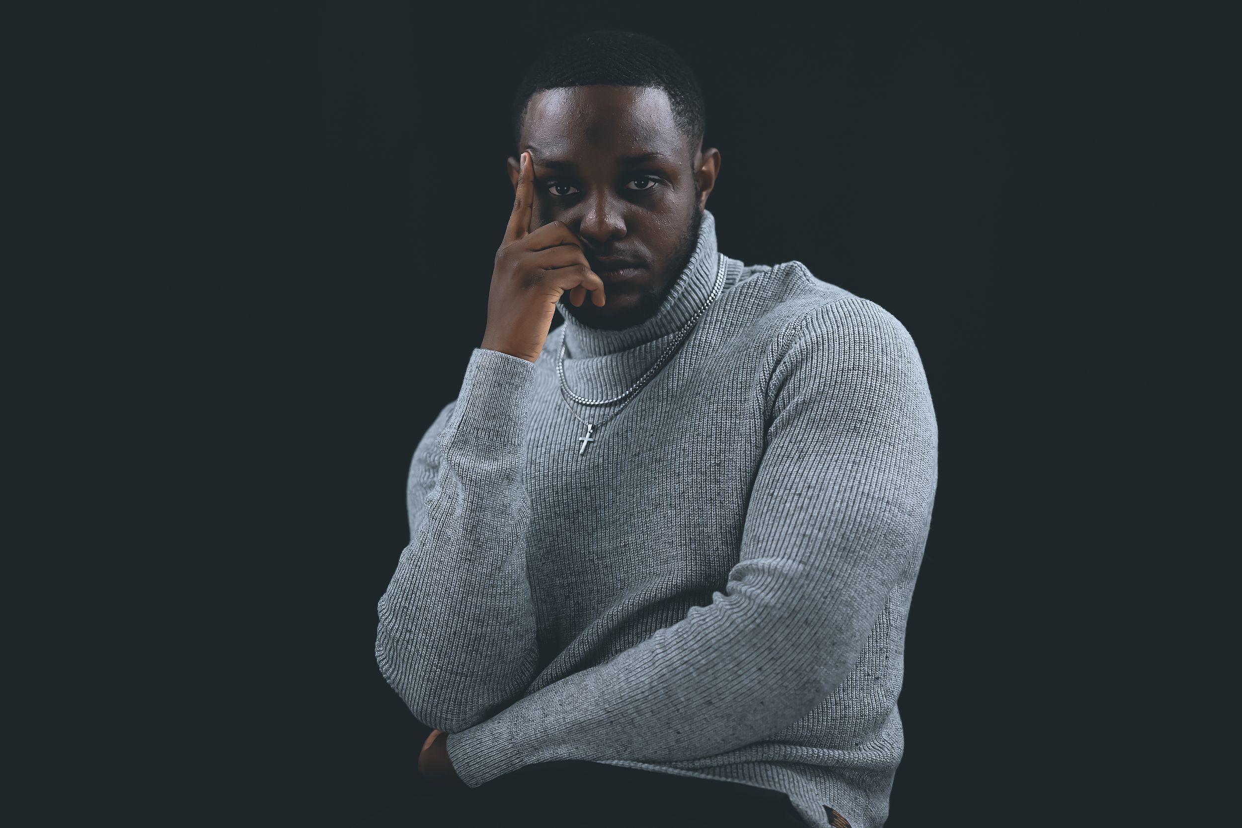 Person wearing a gray sweater stands against dark background in a contemplative pose.