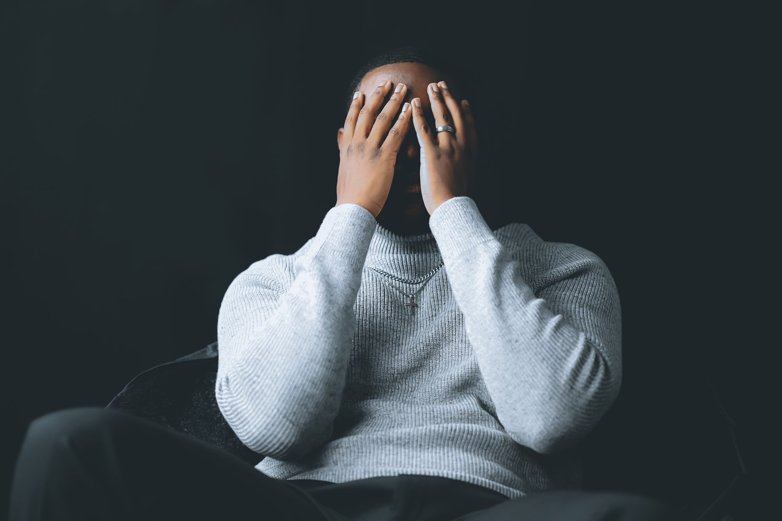 Person in a gray sweater sitting with hands covering their face against dark background.