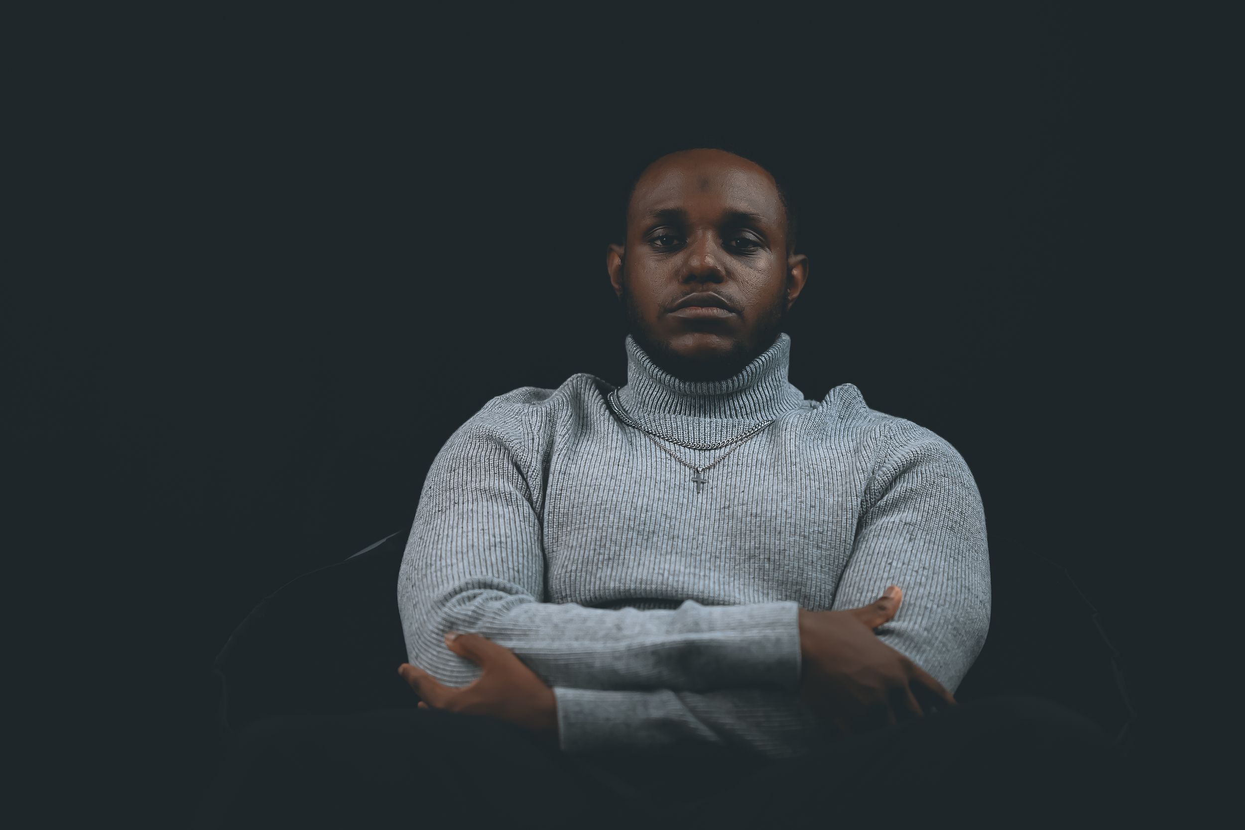 Individual wearing a gray turtleneck sweater poses with crossed arms against dark background.