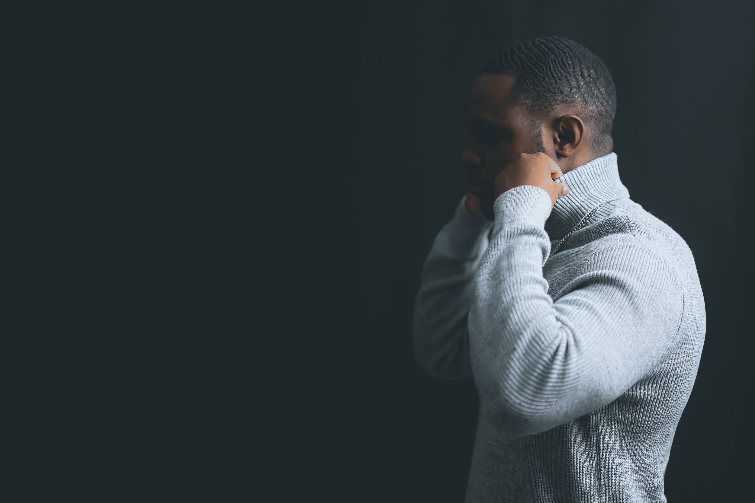 Person in gray sweater holding head in hands against dark background showing signs of distress or sadness.