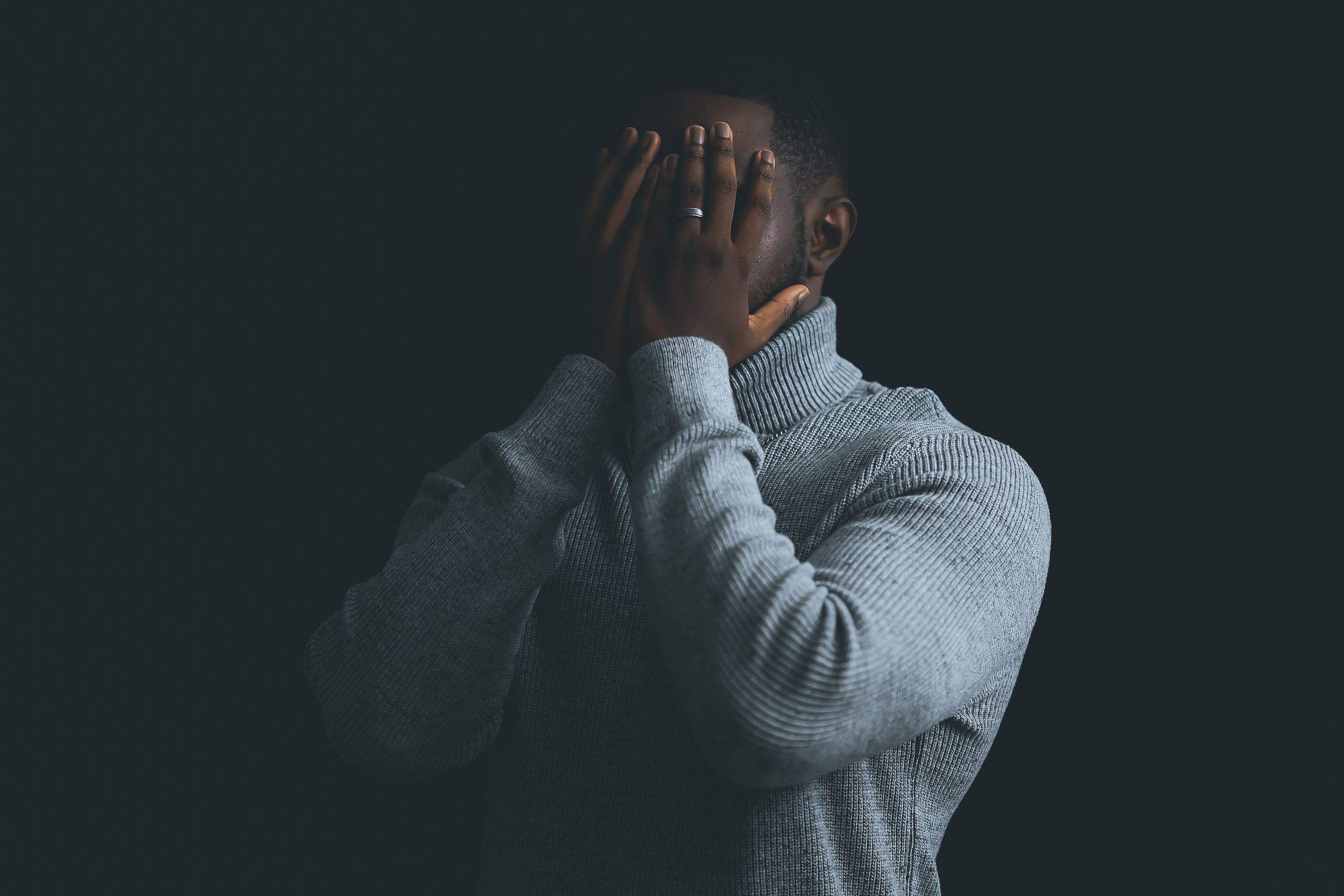 Person in gray sweater covering face with hands against dark background in dramatic lighting.