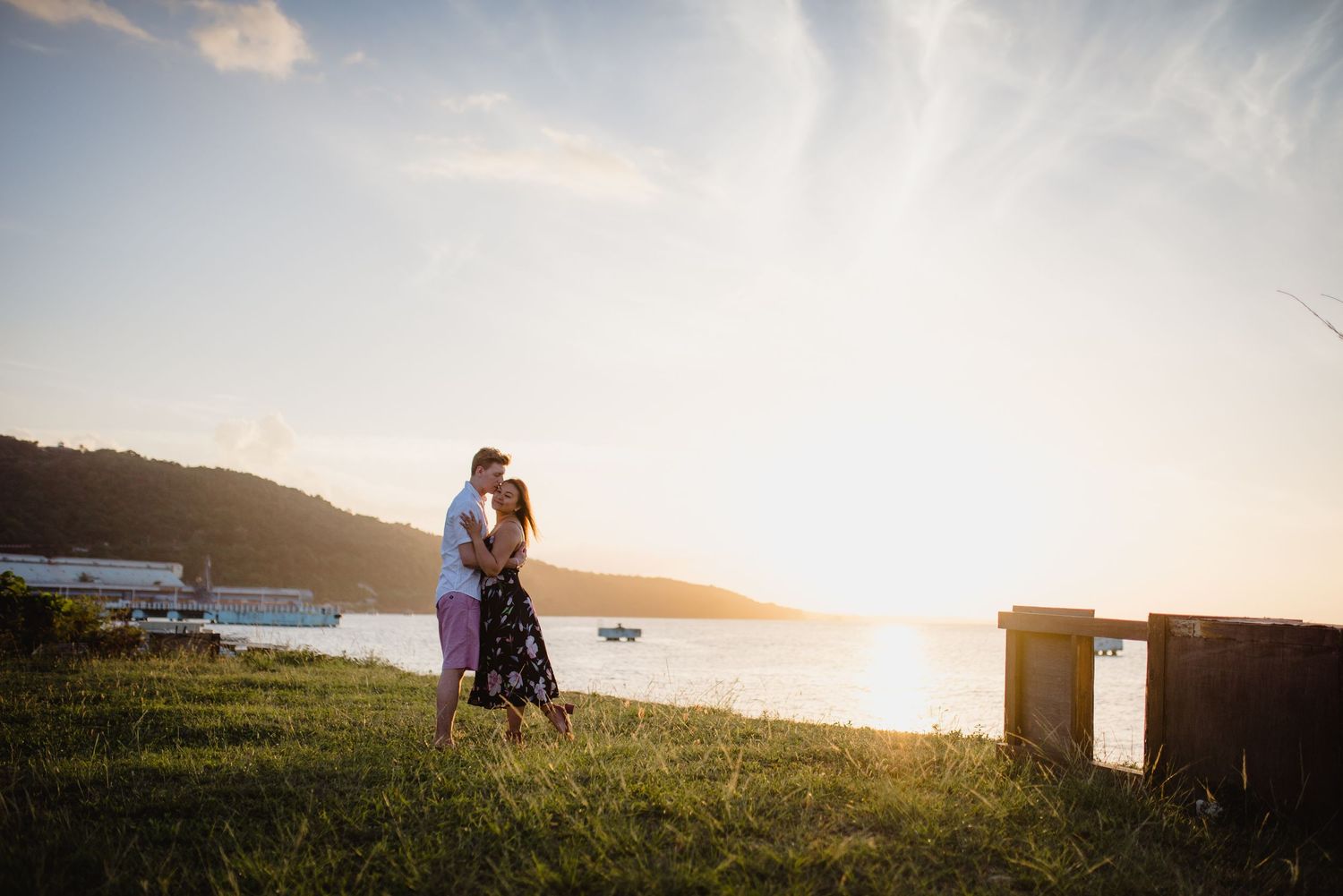 A loving couple stands silhouetted against a vibrant sunset on a grassy hilltop with mountains in the background.
