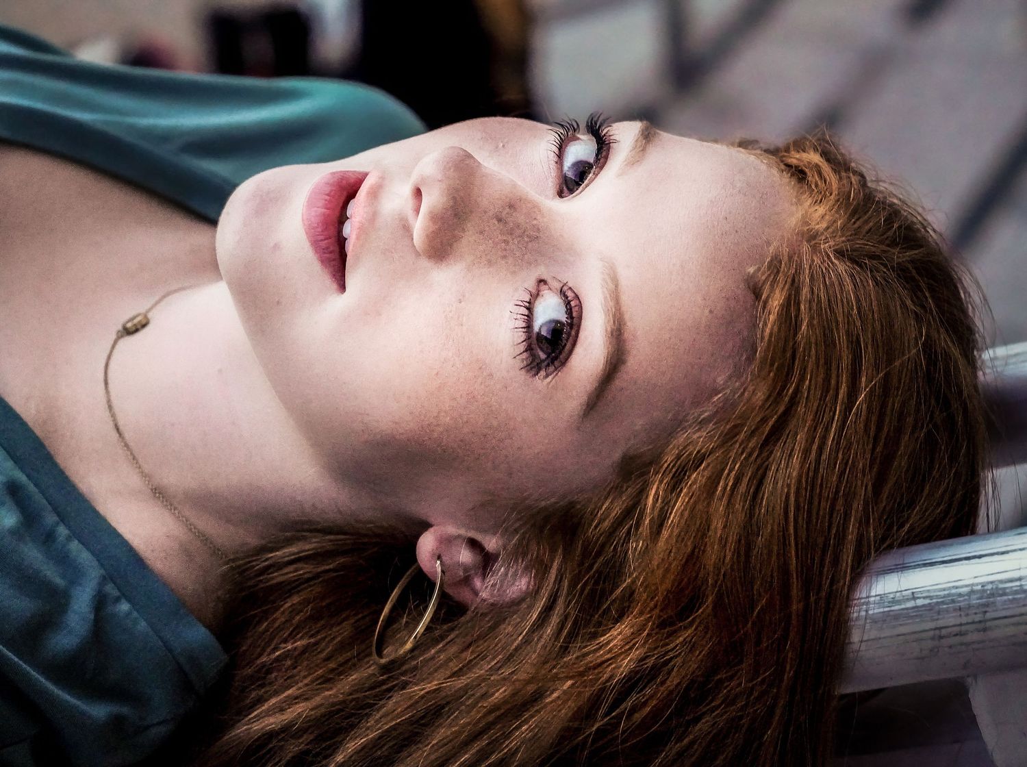 Young woman with red hair and freckles lying back, gazing upward with a contemplative expression outdoors.