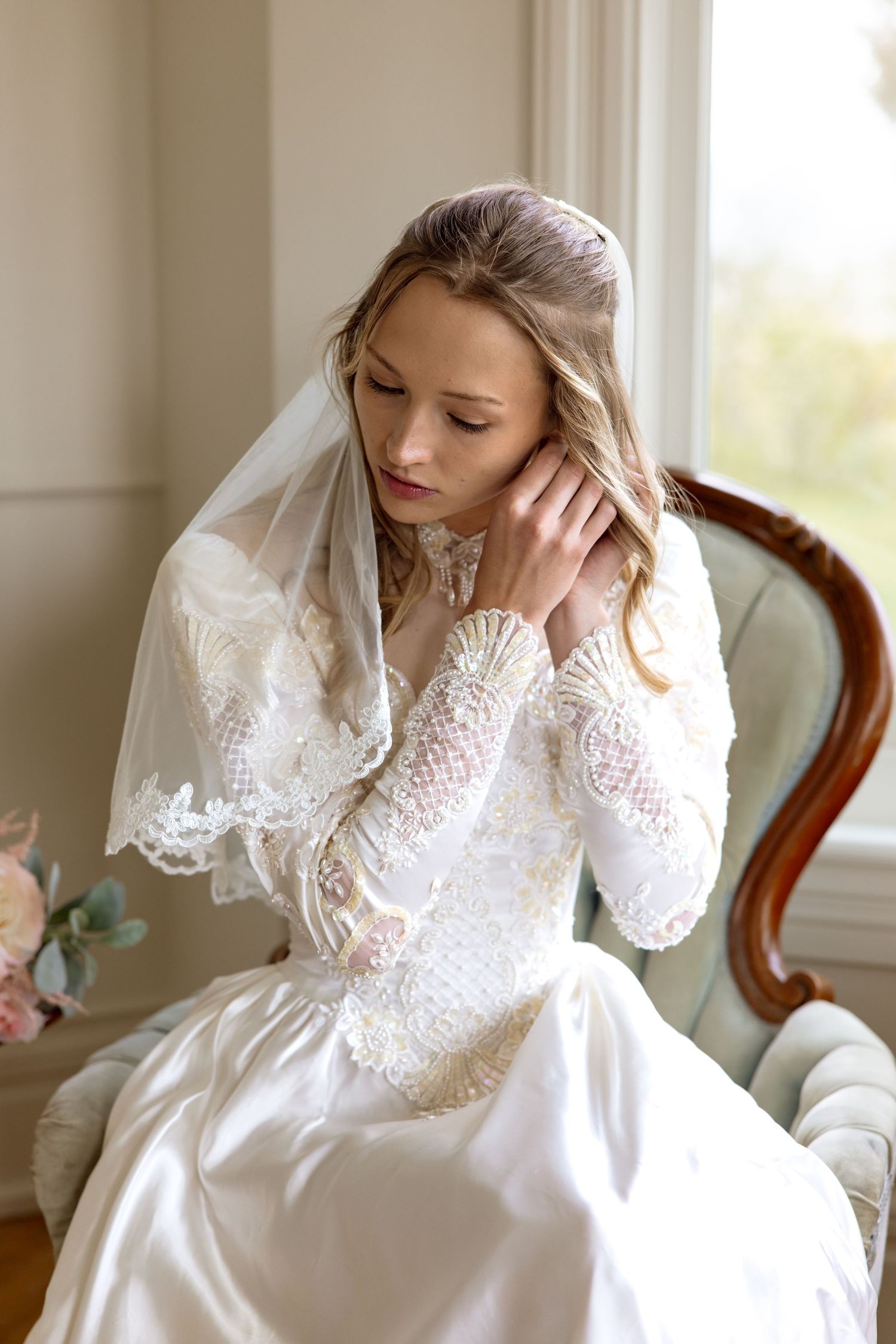 A bride in a vintage-style white lace gown with long sleeves sits thoughtfully by a window in soft natural light.