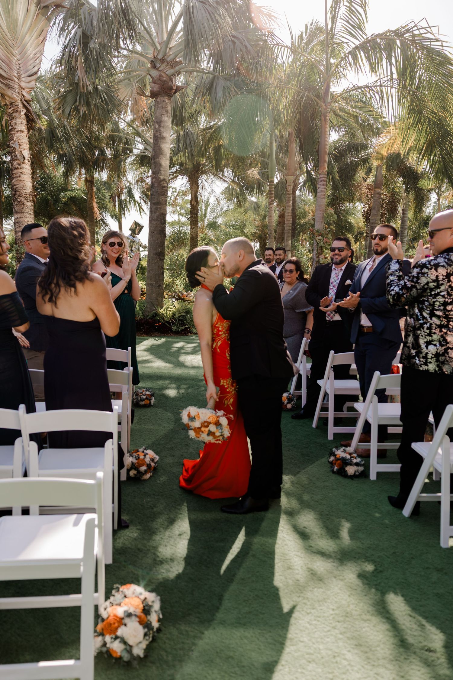 Romantic outdoor wedding ceremony with guests gathered under palm trees on a green carpet walkway with white chairs.