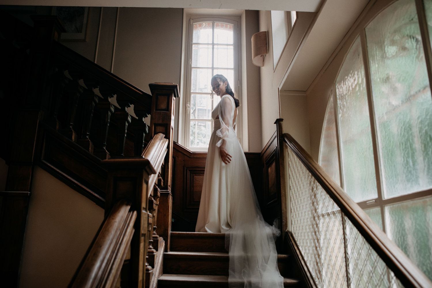 Bride in a flowing white gown standing on wooden staircase by a large window.
