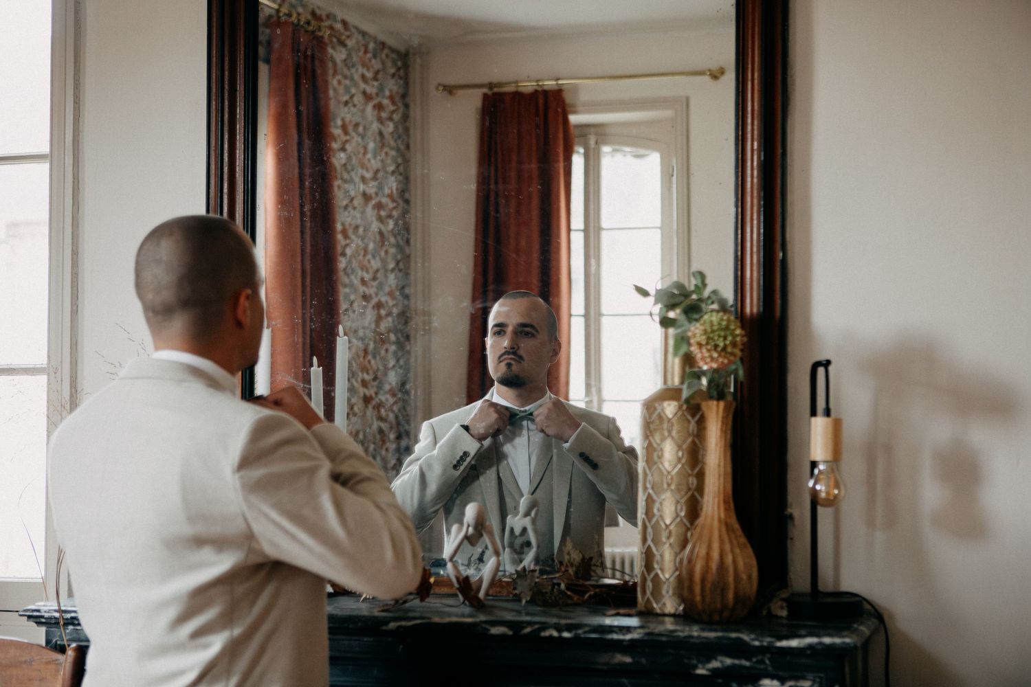 Groom in a beige suit adjusting his tie while looking in an ornate mirror.