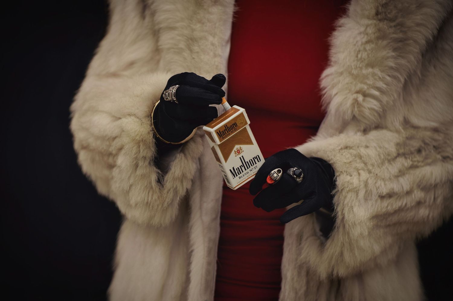 Close-up of a person in a fur coat holding a Marlboro cigarette pack, wearing black gloves and rings. Photo by Carolina Waldroff.
