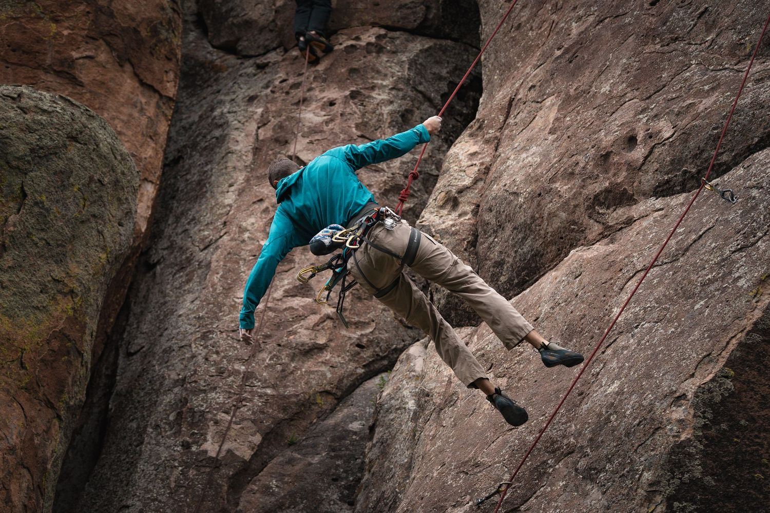 Rock climber in turquoise jacket scales steep cliff face while secured by climbing gear and ropes.