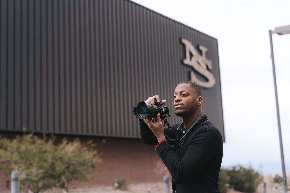 A photographer in a black jacket holds up a camera to take photos outside a building with an NS logo.