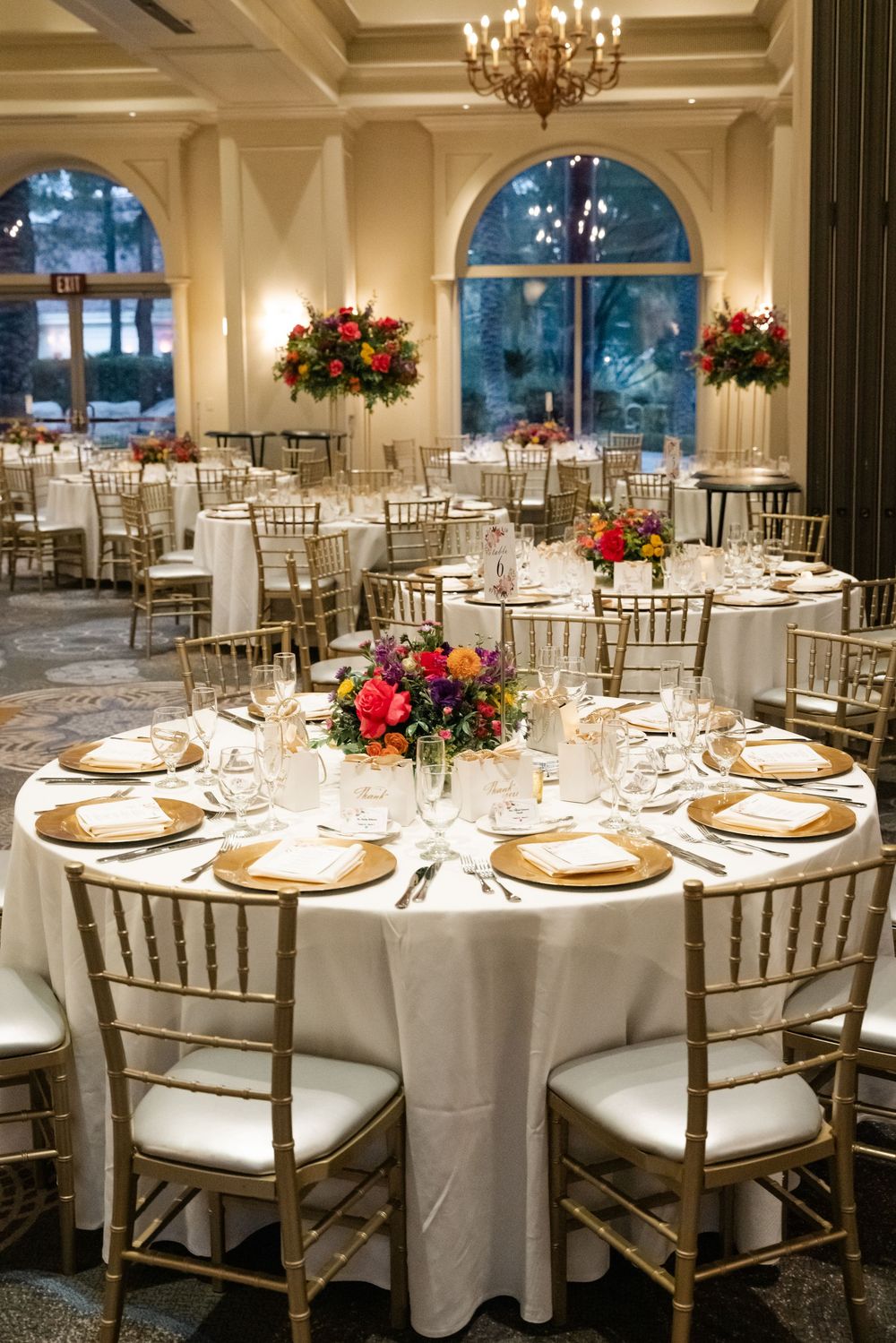 Elegant ballroom wedding reception with round tables, gold chiavari chairs, crystal chandelier and vibrant floral centerpieces.