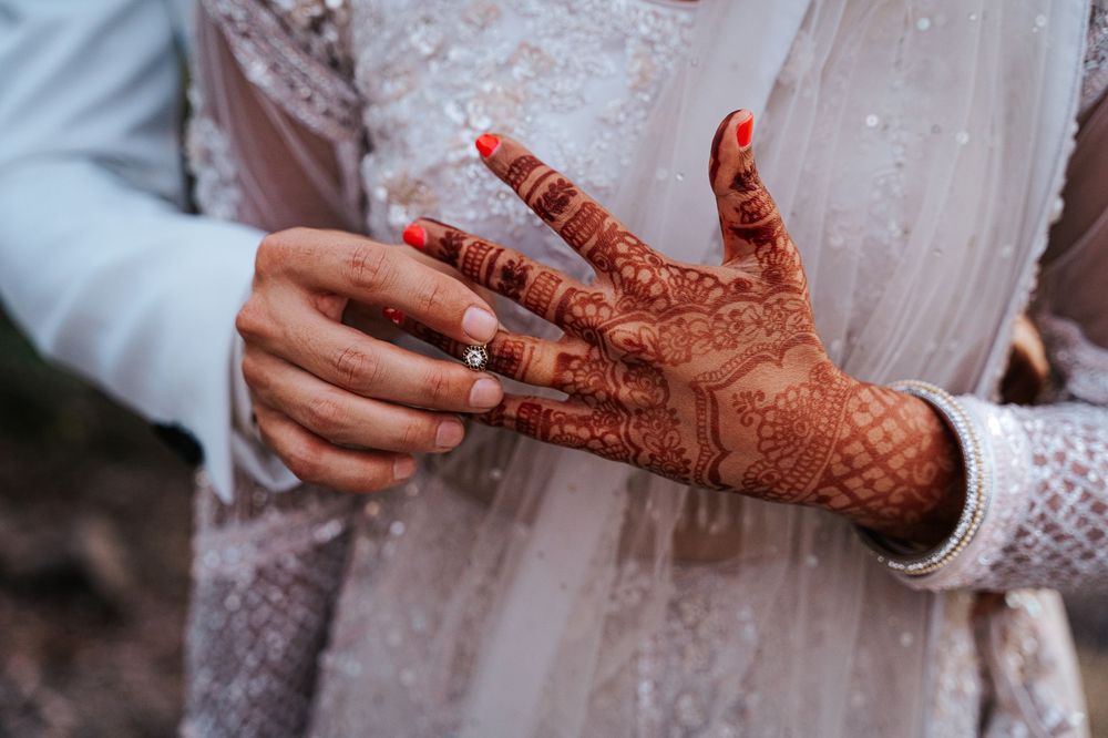 Close up detail of intricate traditional henna design on hands with white wedding attire visible in background.