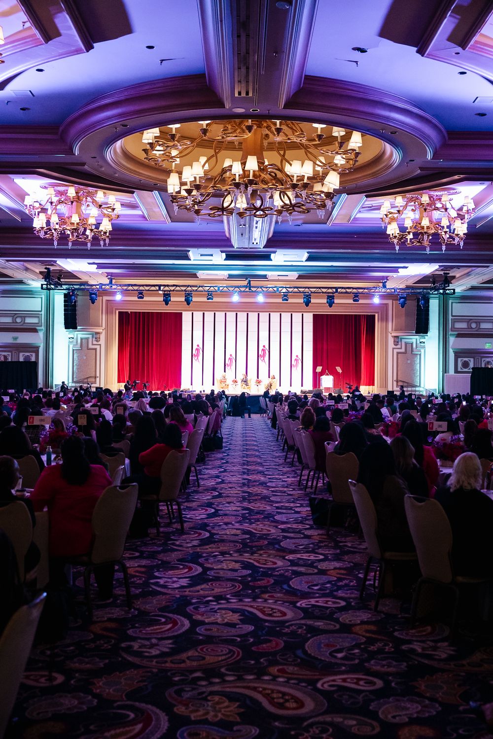 Grand ballroom with ornate chandeliers and red stage curtains filled with attendees seated in rows on patterned carpet.