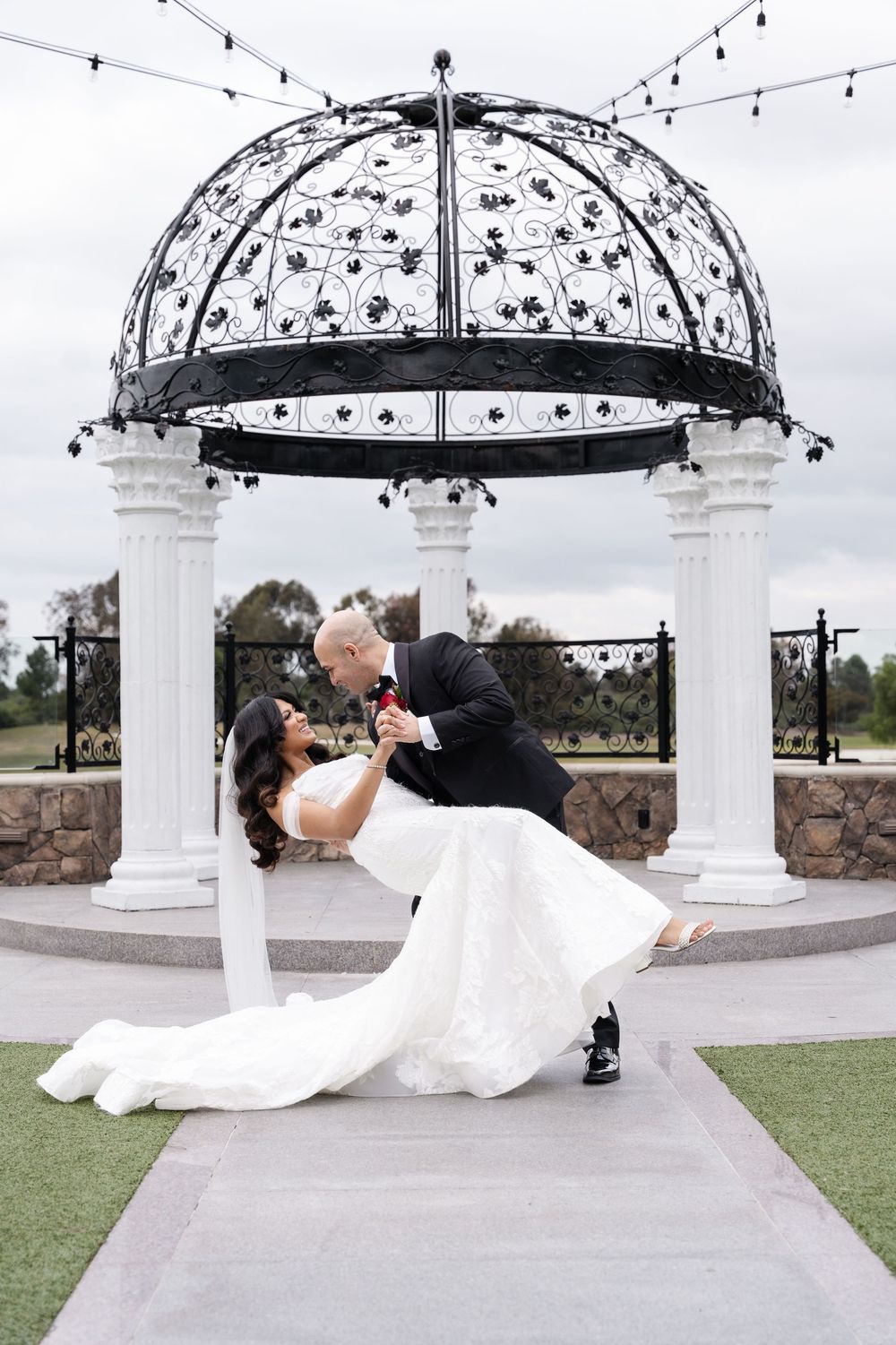 Romantic couple shares a dip kiss under an elegant white gazebo with ornate metal dome and hanging string lights outdoors.