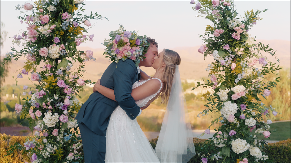 A romantic outdoor wedding ceremony with two people embracing under floral arches at sunset.