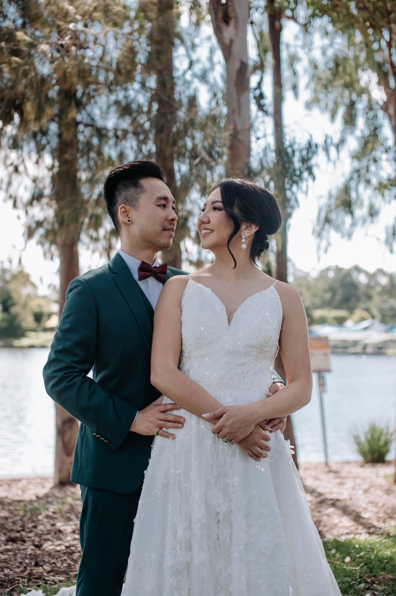 A newlywed couple shares a romantic moment by a lake, with eucalyptus trees in the background.