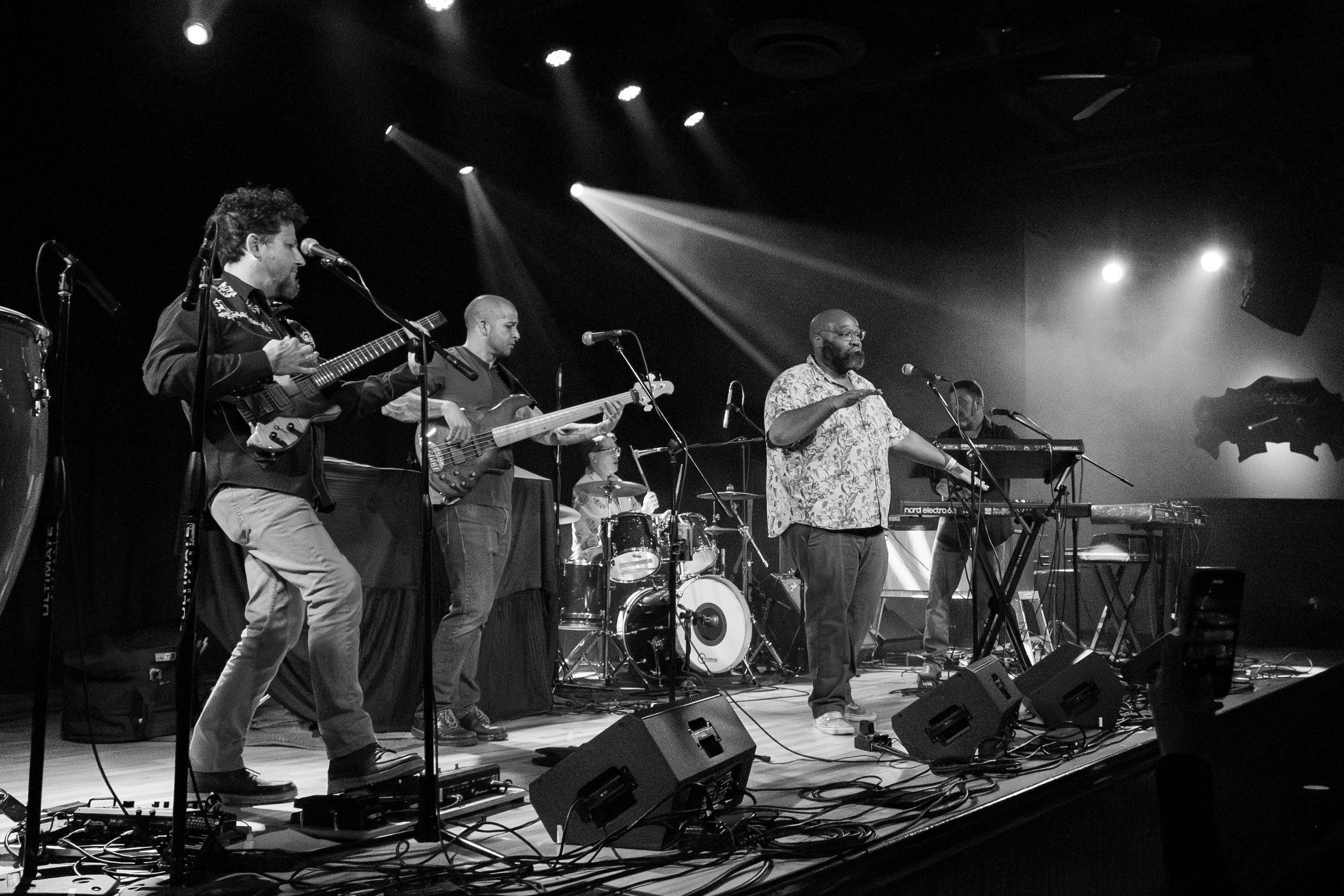 Wide angle black and white shot of full band performing live on stage with multiple instruments and equipment.
