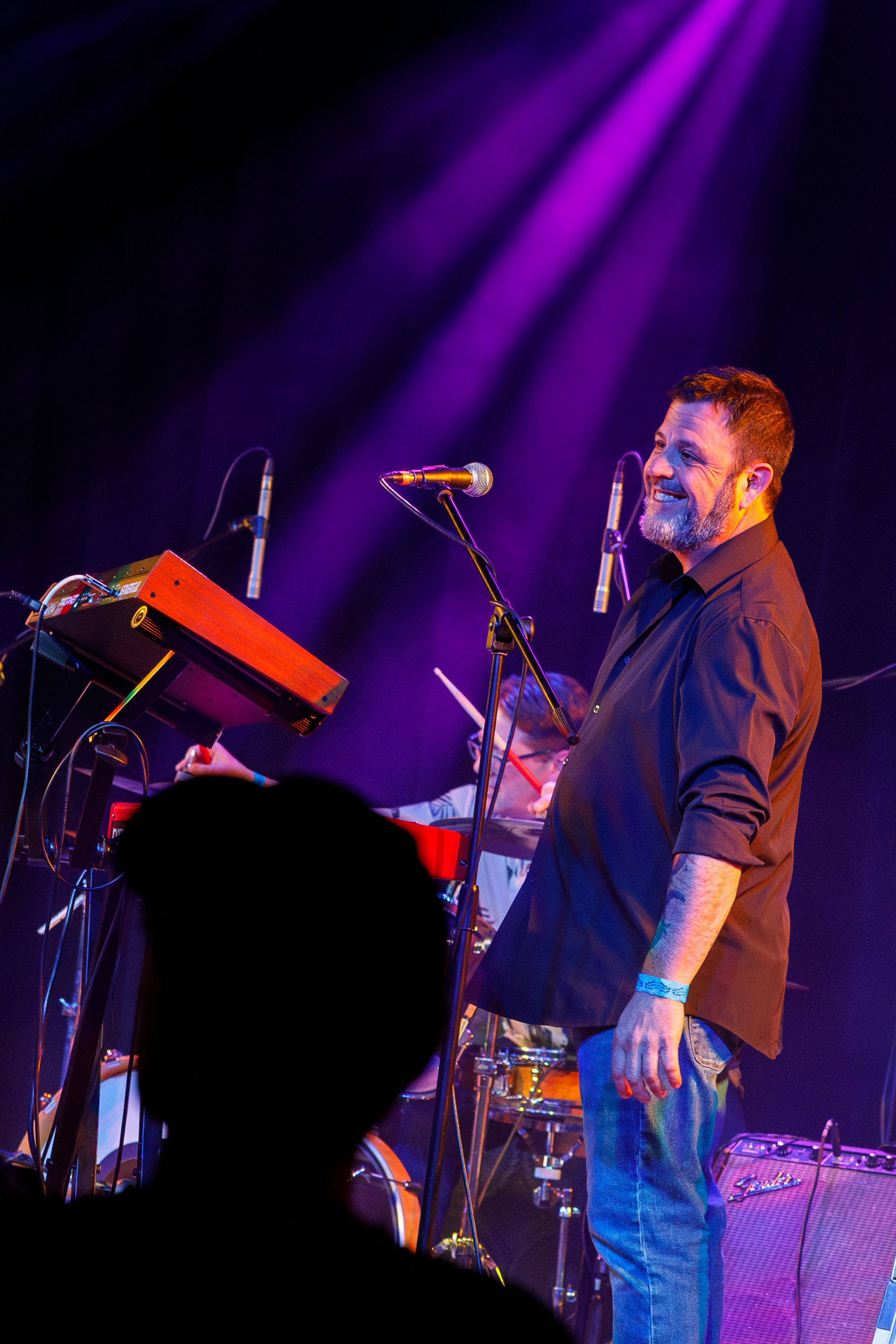 A musician performs on stage under dramatic purple lighting with keyboard equipment and microphones visible.