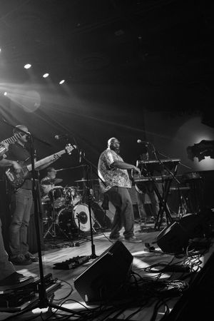 Black and white concert photo showing band members performing on stage with instruments and stage lighting.