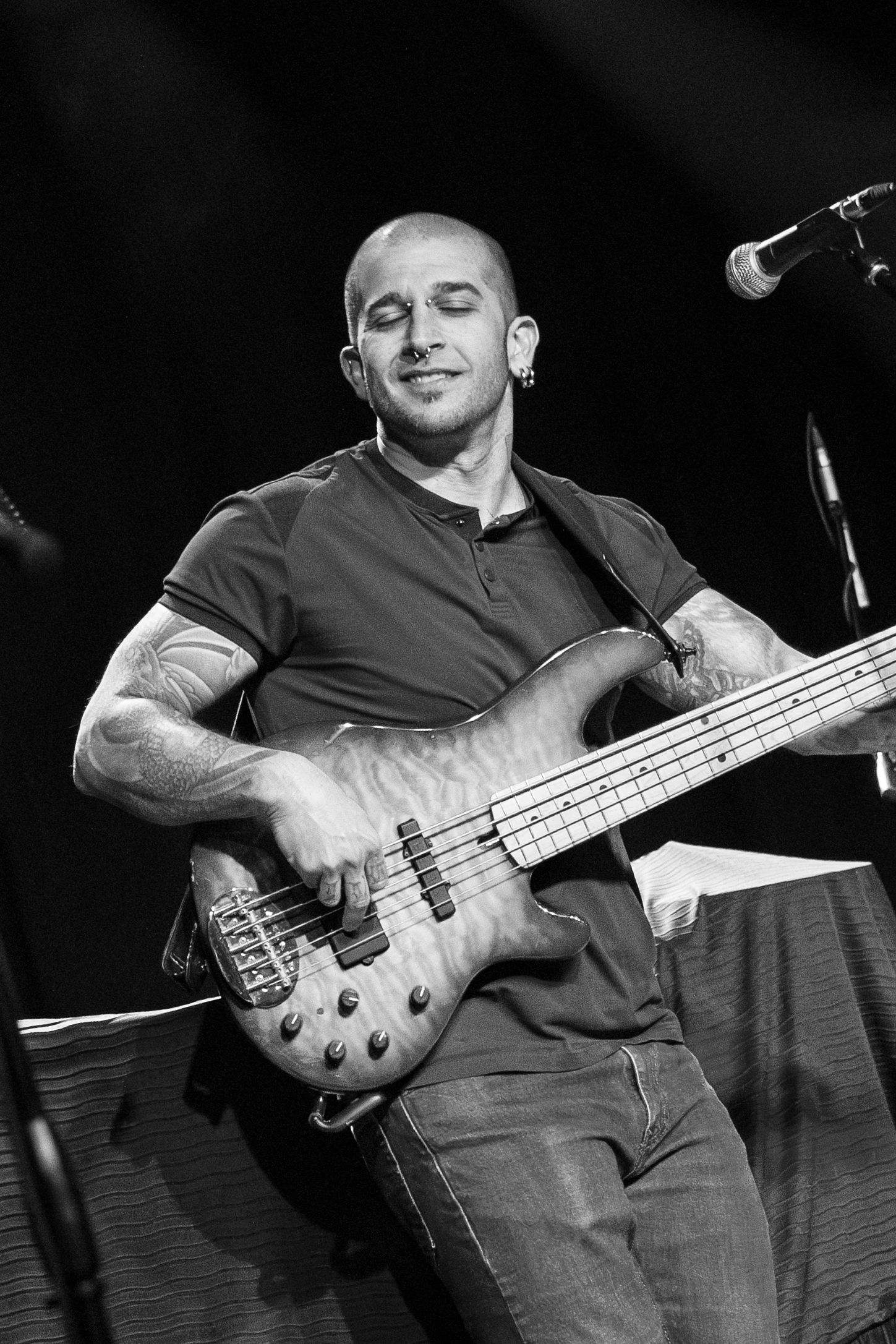 A bass guitarist performs on stage in black and white, displaying intricate tattoo work while playing a deluxe instrument.