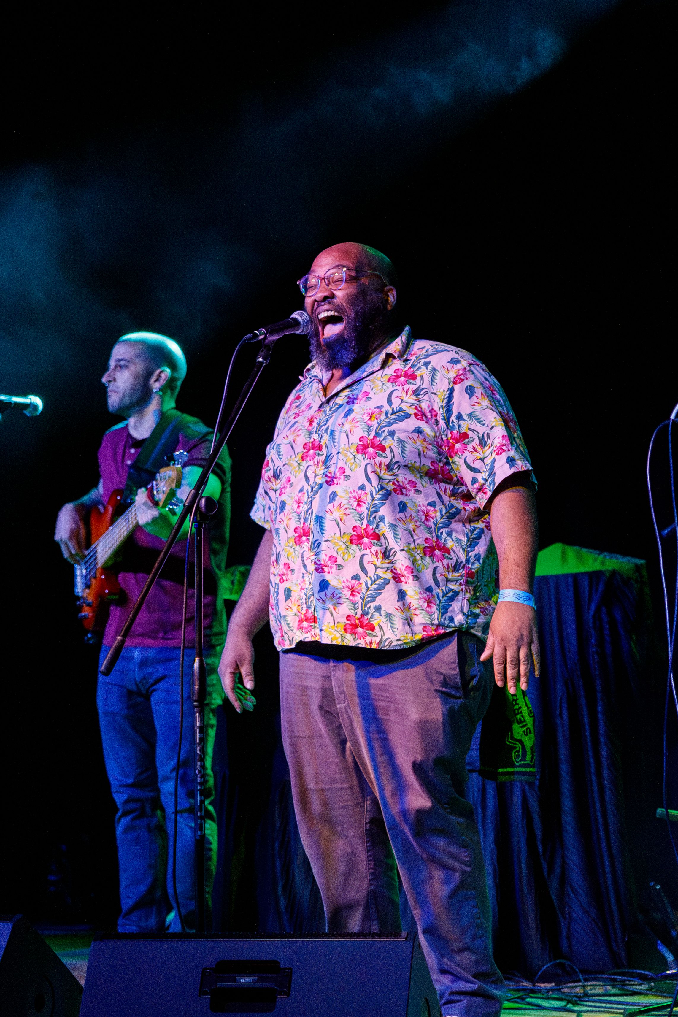 Performer in colorful floral shirt passionately singing on stage with guitarist in background during live concert performance.