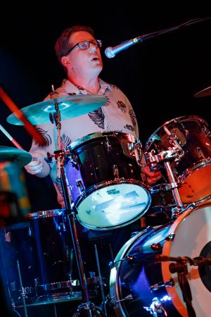 Drummer wearing glasses enthusiastically plays drum kit during dynamic stage performance with blue lighting.