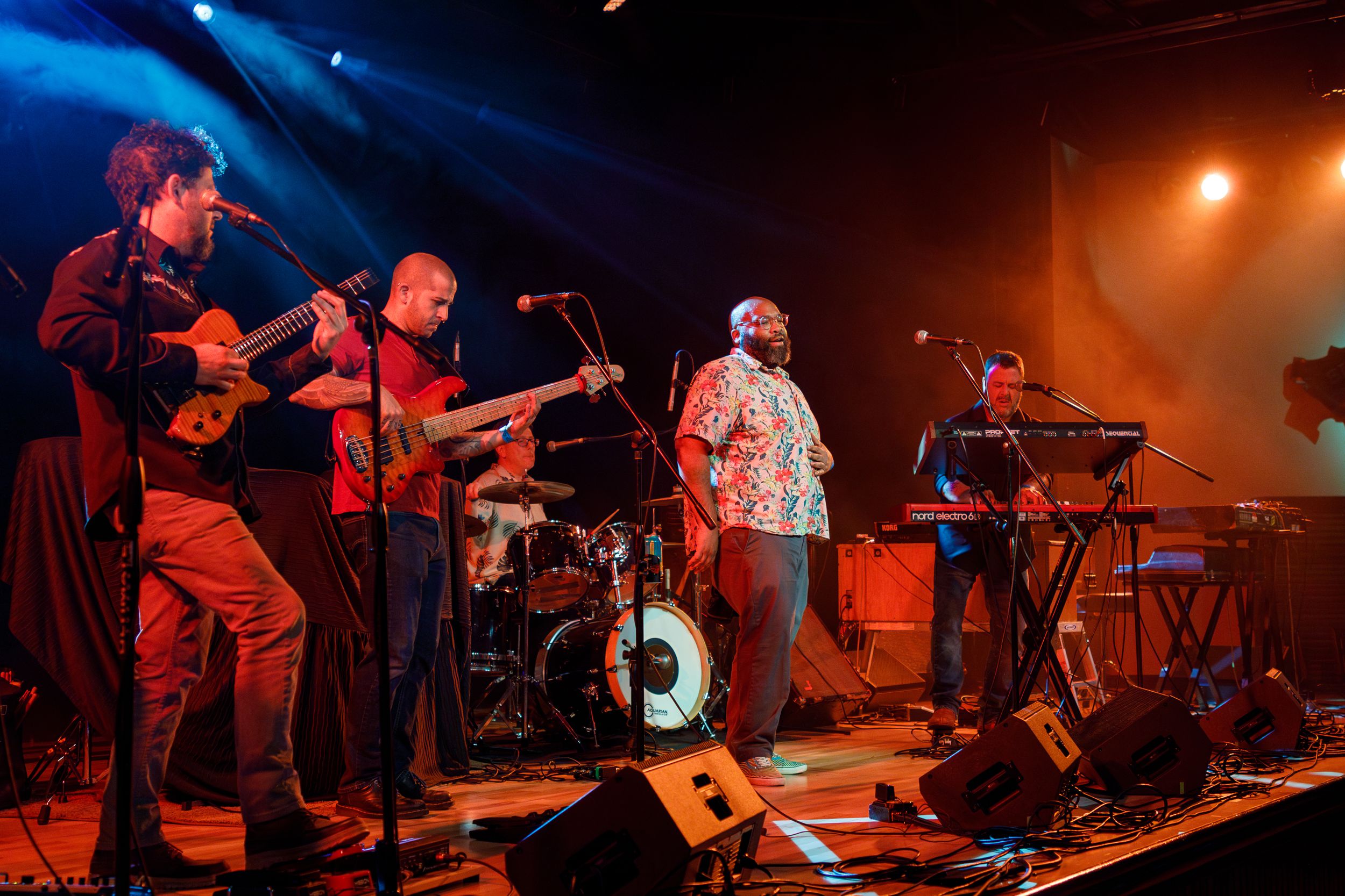 Musicians perform on stage with guitars, drums and keyboards under dramatic blue and orange concert lighting.