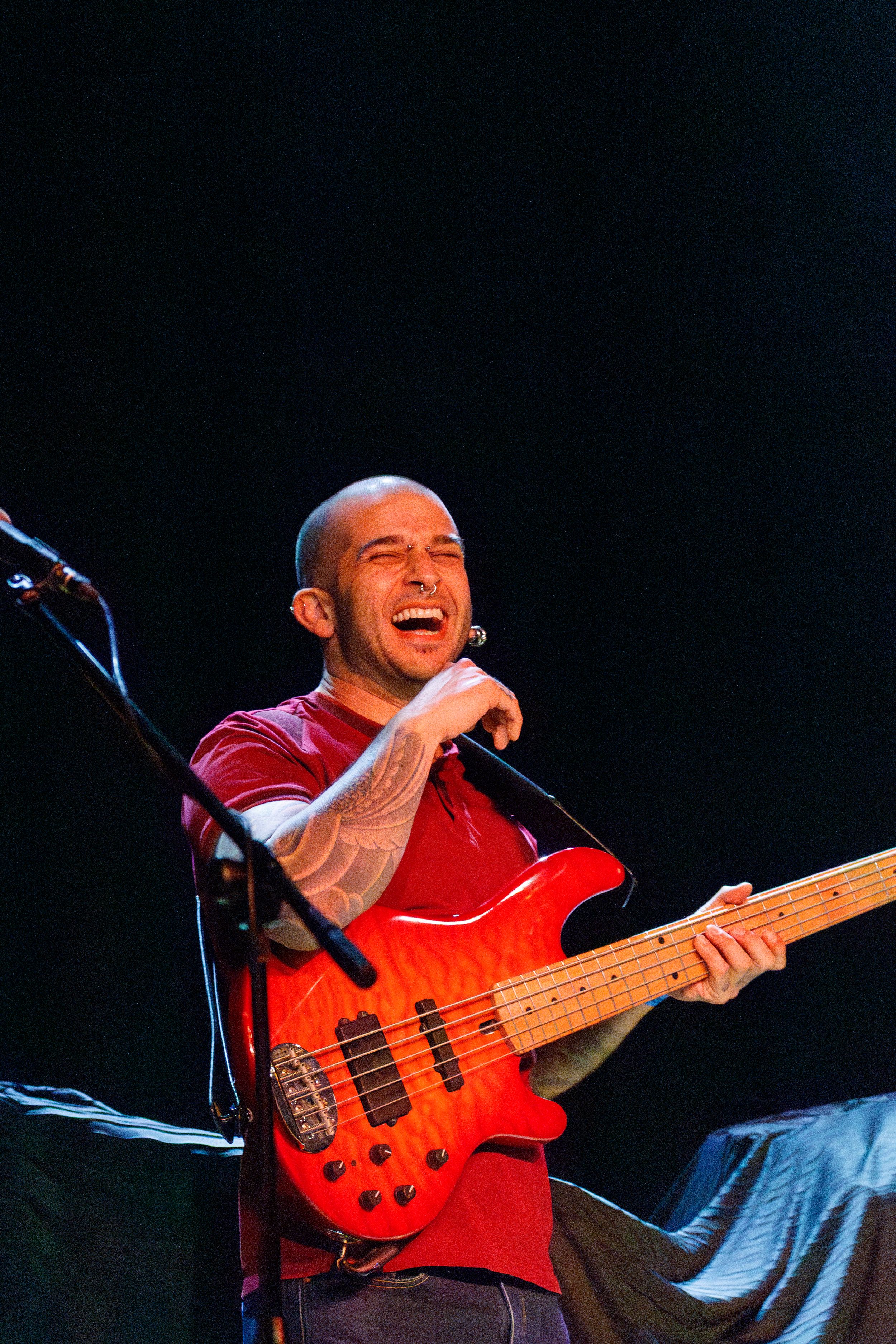 Musician performs with red bass guitar on dark stage, smiling with joy during live performance.