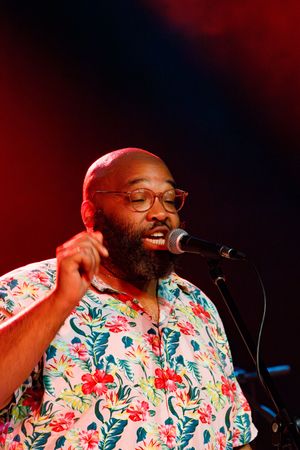 Performer in floral shirt sings passionately into microphone under red stage lighting.