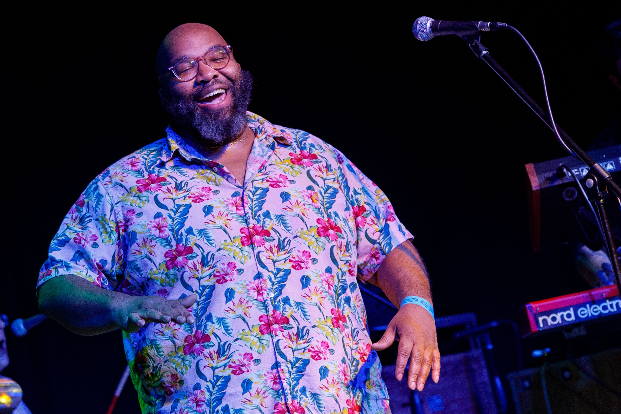 Musician performing on stage wearing a colorful floral print short-sleeve shirt under blue stage lighting.