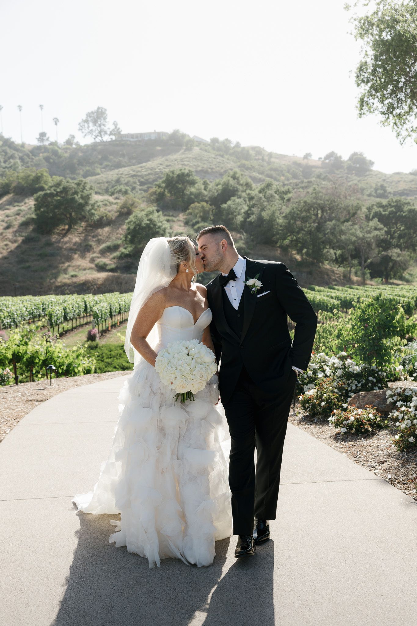 A romantic wedding couple shares a tender moment on a path surrounded by vineyards and rolling hills on a sunny day.