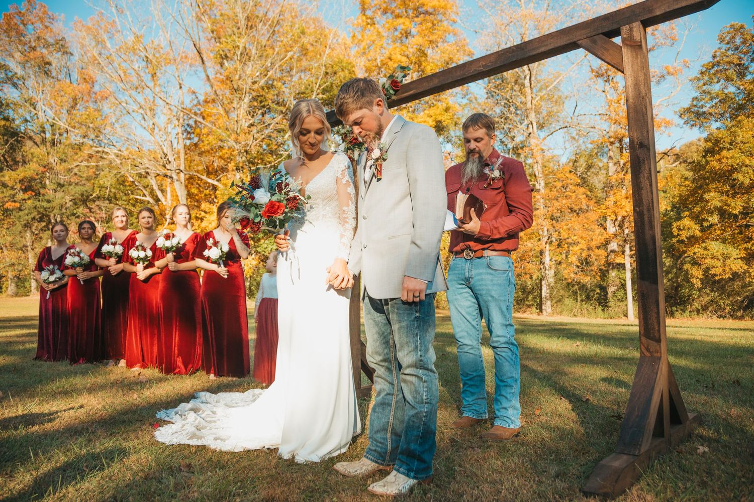 Wedding ceremony guests gather outdoors under autumn trees with rustic wooden decor.