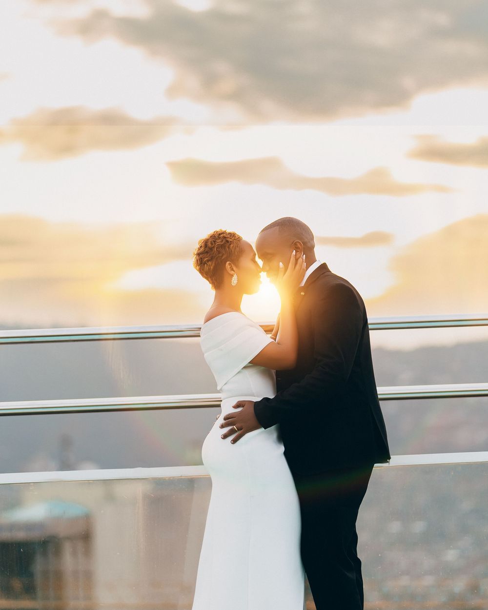 Two figures in elegant wedding attire embrace on a balcony during golden sunset with city backdrop.