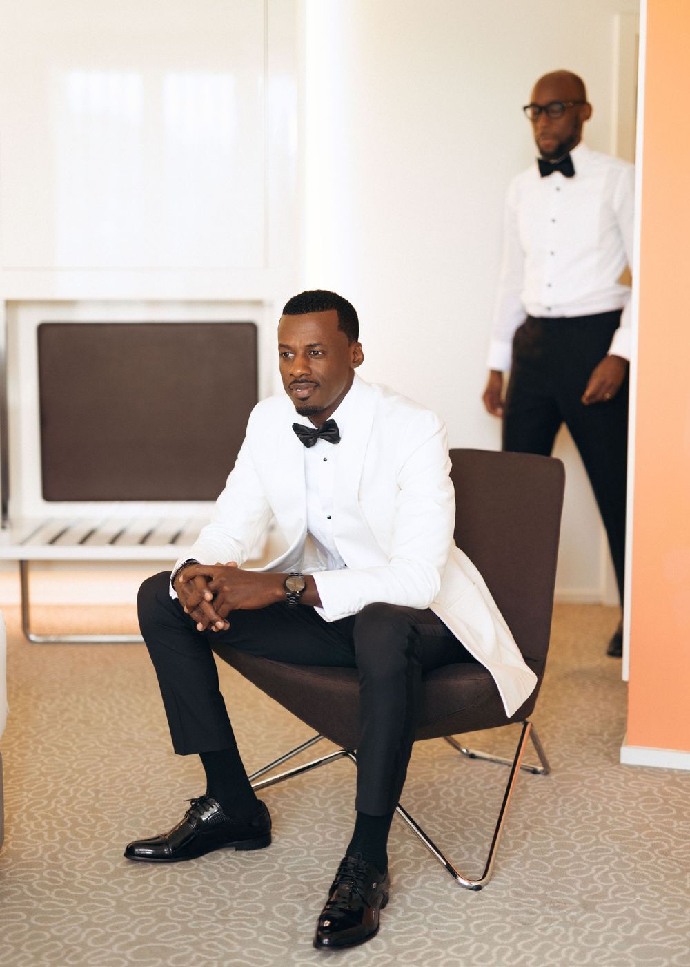 A person in a crisp white tuxedo jacket and bow tie sits elegantly on a brown modern chair in a minimalist setting.