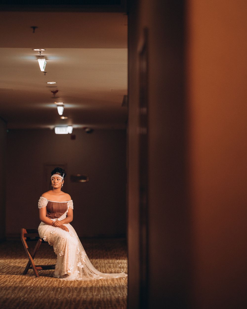 Someone in an elegant off-shoulder gown poses on a wooden chair in a dimly lit hotel hallway.