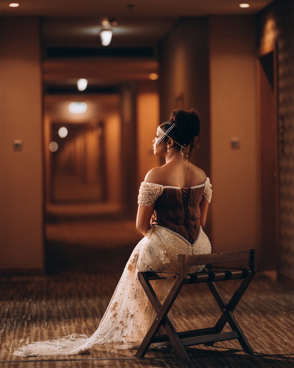 Person in elegant off-shoulder lace wedding gown sits on director's chair in warm-lit hallway with dramatic lighting.