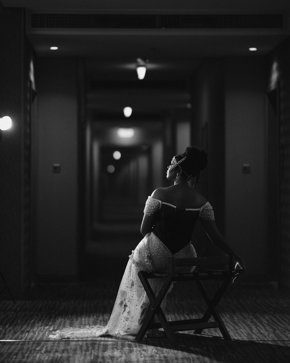 A person in a dress sits on a folding chair in a dimly lit hallway with overhead lights creating a moody atmosphere.