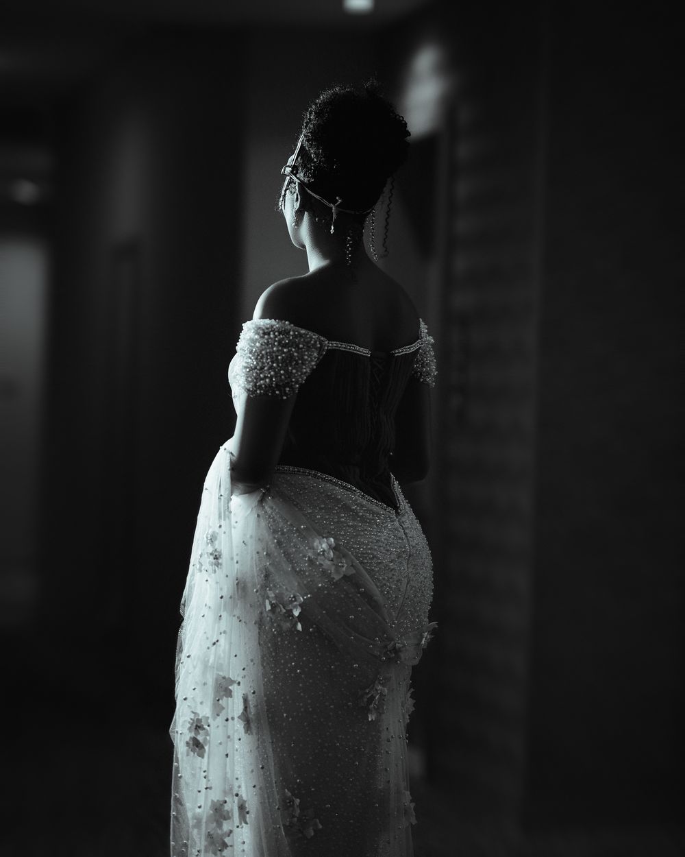 Black and white silhouette of a bride in an elegant off-shoulder lace wedding gown captured from behind in dramatic lighting.