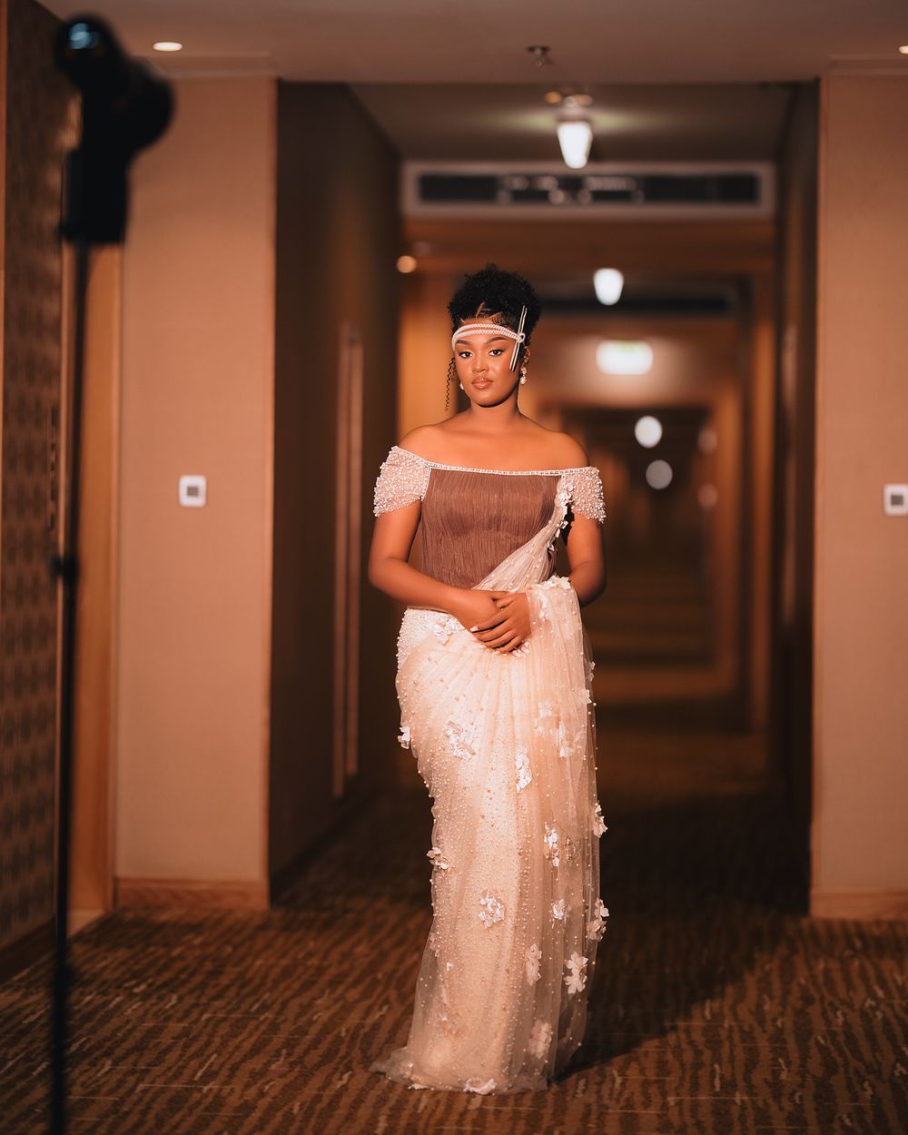 A person poses elegantly in a hallway wearing an off-shoulder cream gown adorned with delicate floral appliques.
