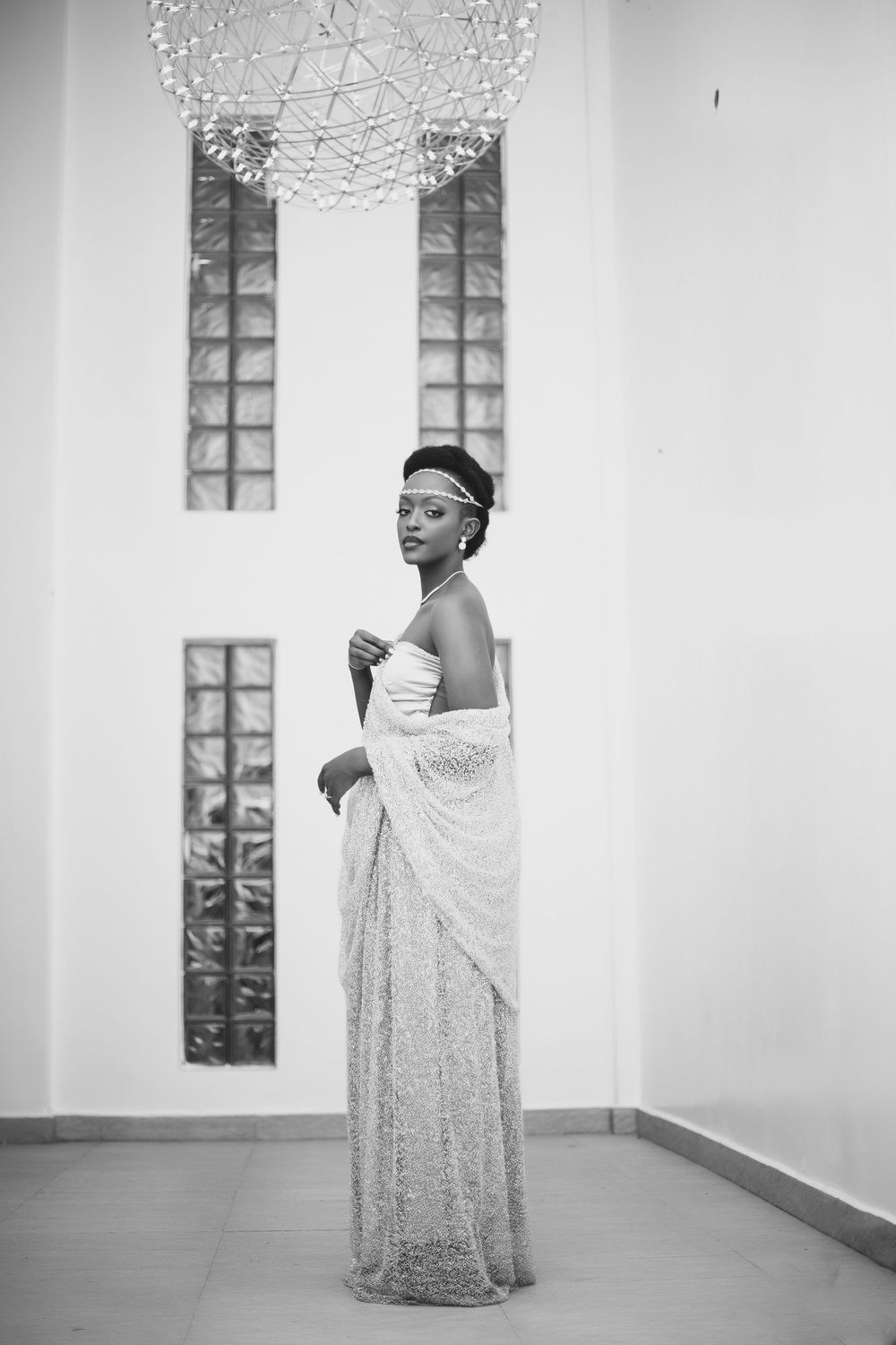 Elegant black and white photograph of wedding gown featuring strapless bodice and flowing lace wrap under crystal chandelier.