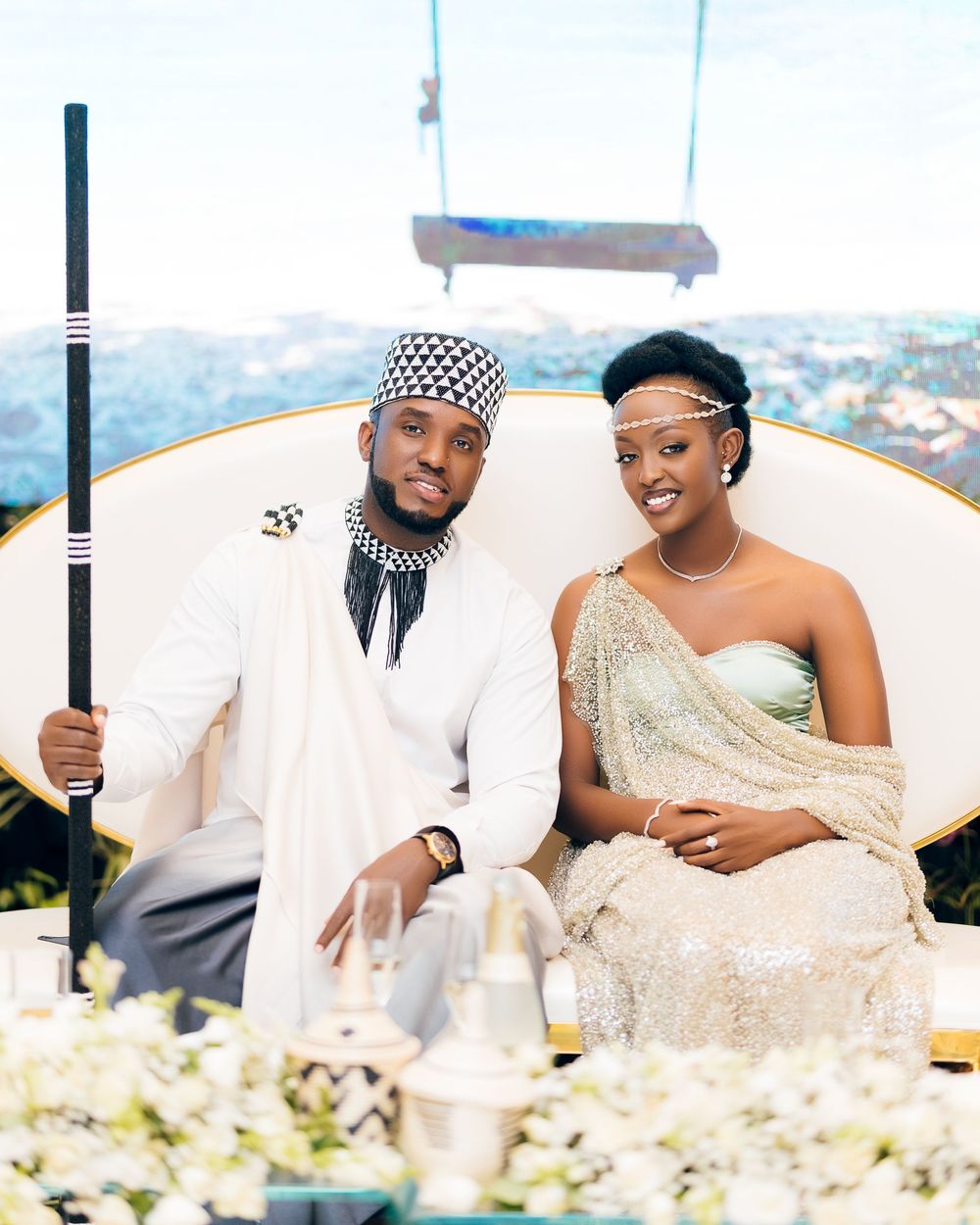 Well-dressed couple smiling while sitting together at elegant outdoor reception with floral decorations.