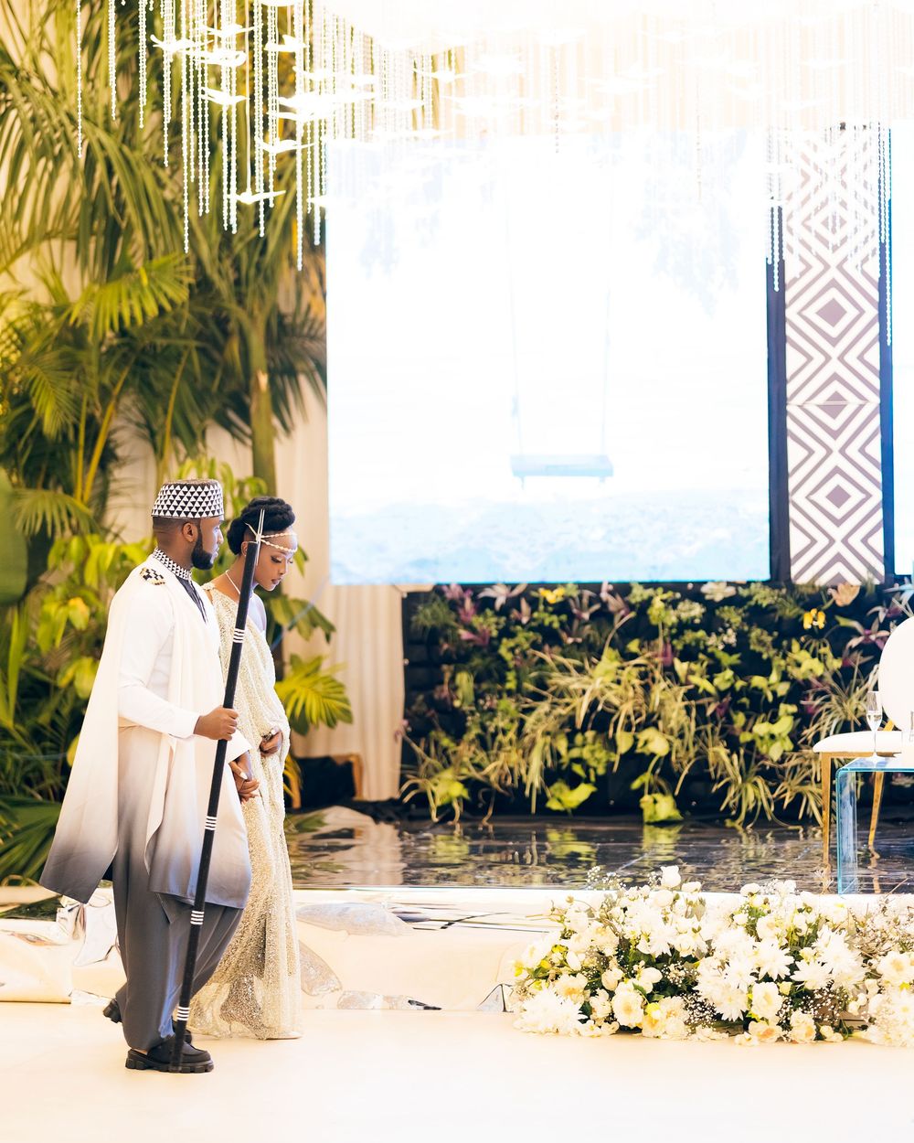 A couple in traditional white and gray wedding attire stands near tropical plants and floral decorations at an outdoor venue.