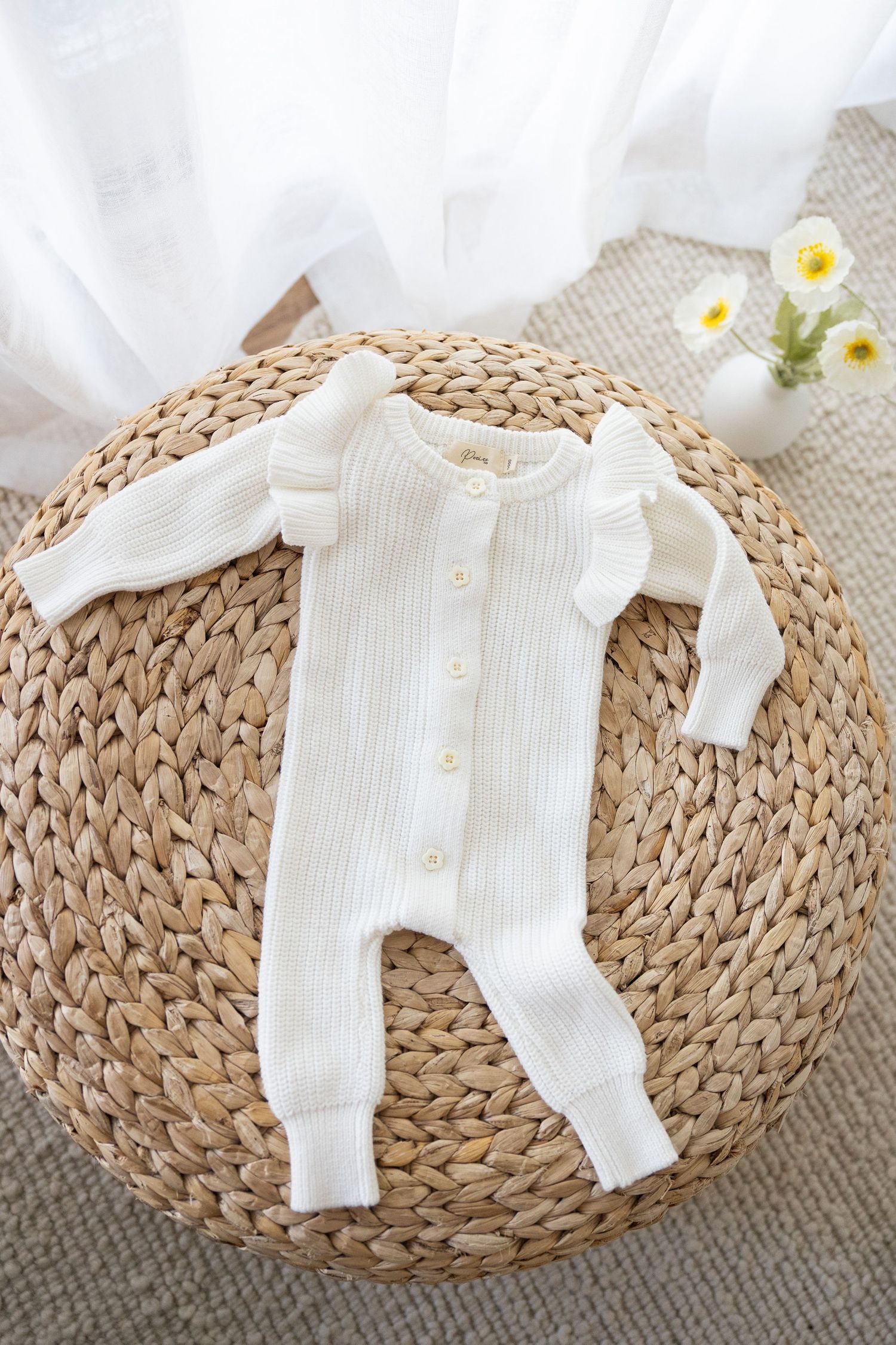 White knit baby romper with ruffled sleeves displayed on circular woven rattan mat with white flowers in background.