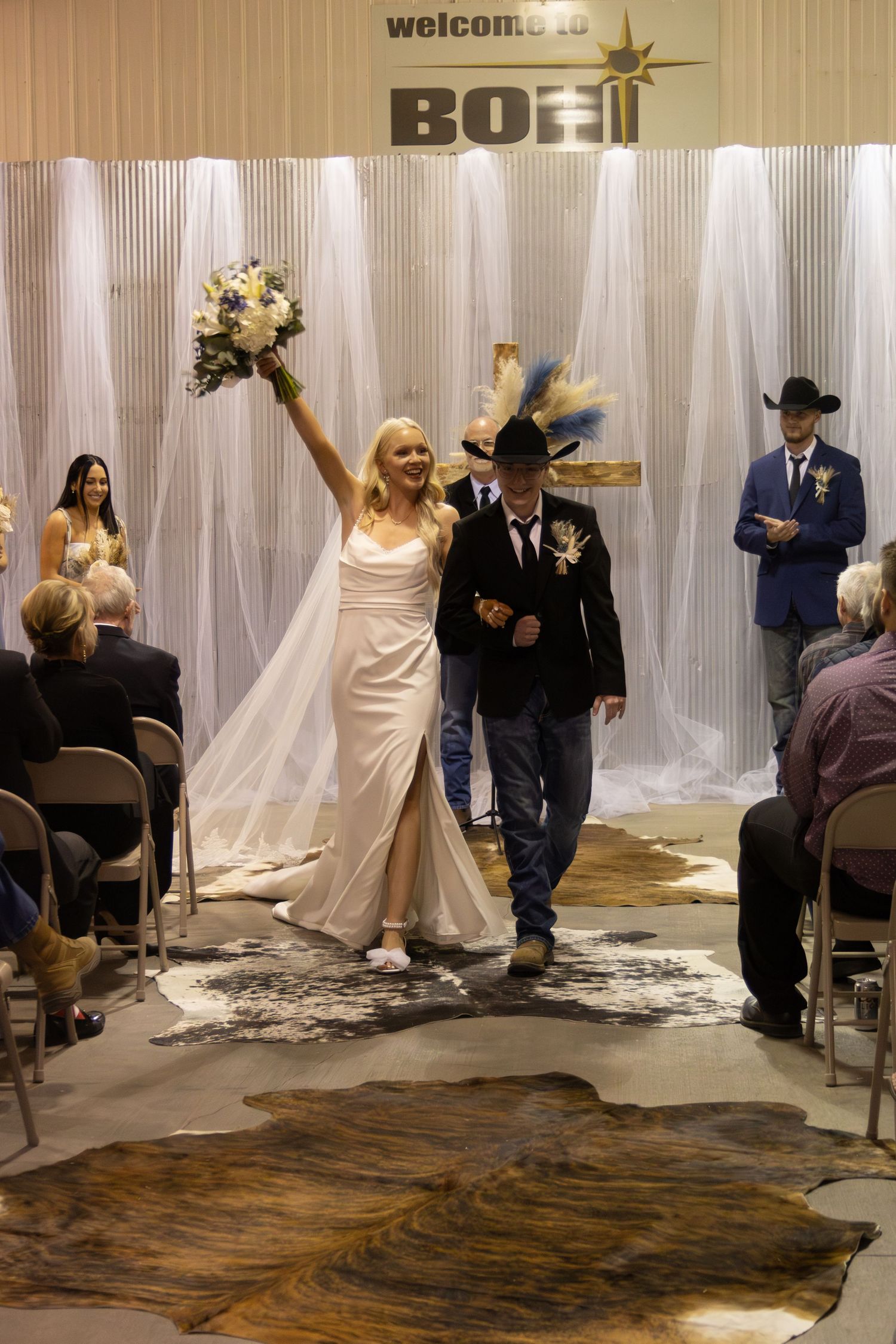 Newlywed couple celebrates while walking down aisle at rustic indoor wedding ceremony with cowhide rug and draped backdrop.