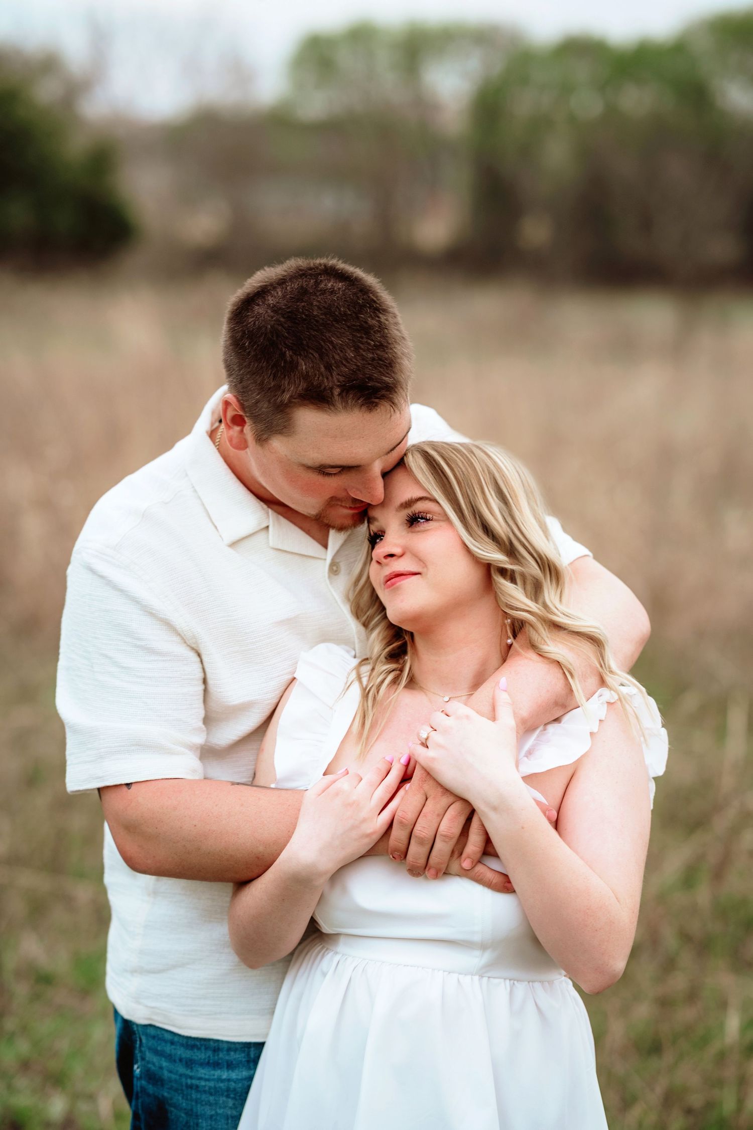 Romantic couple embraces in field wearing coordinating white outfits during golden hour photo session.
