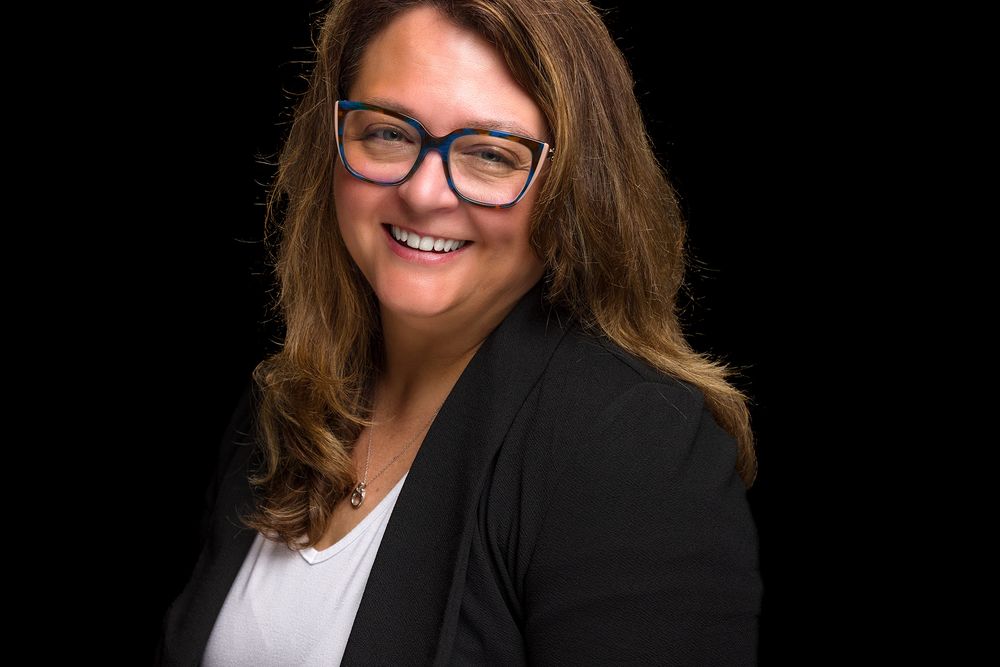 Professional business portrait of a person wearing glasses, a black blazer and white top against a dark background.