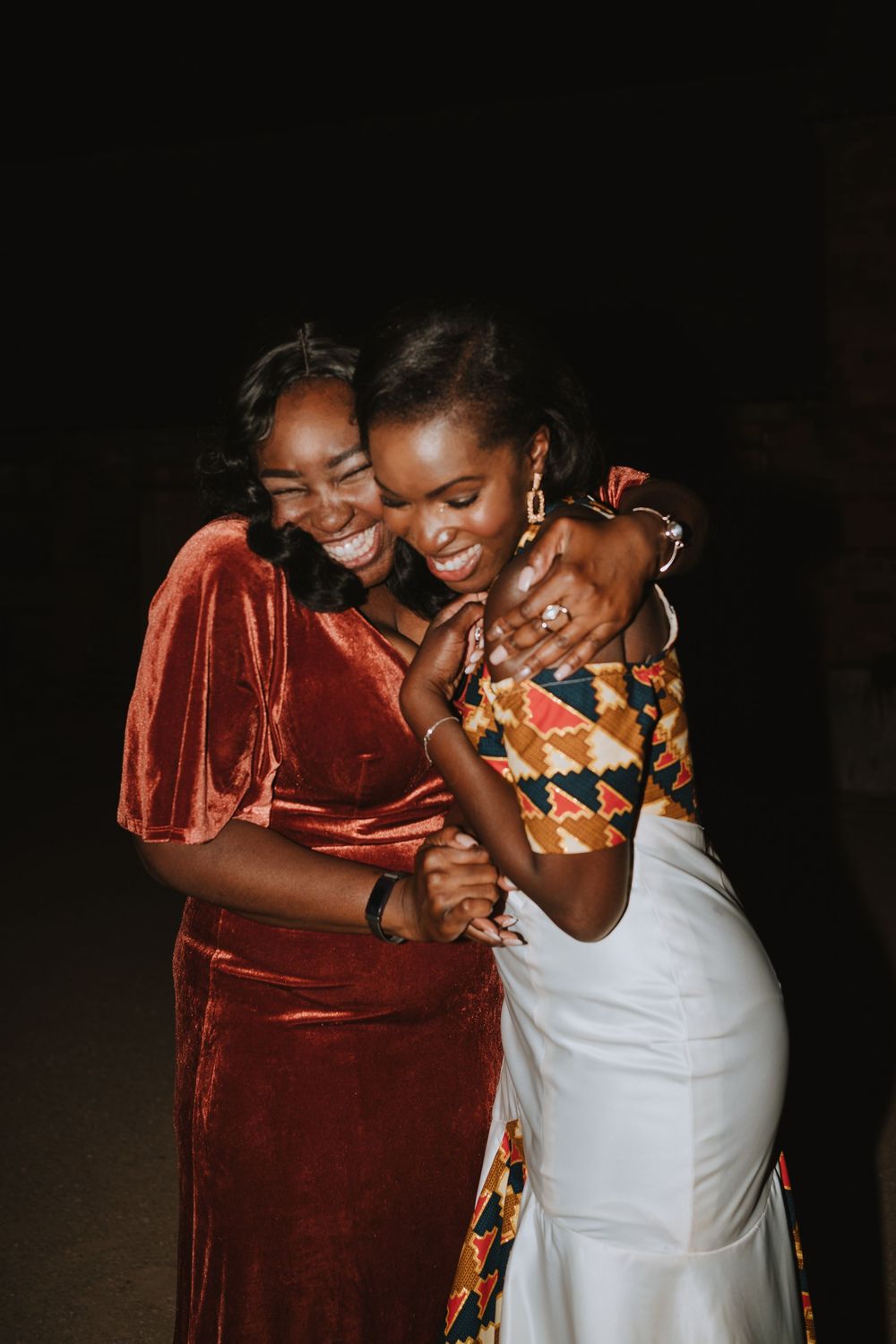Two women share a joyful embrace while wearing an elegant rust velvet dress and white dress with African print details.